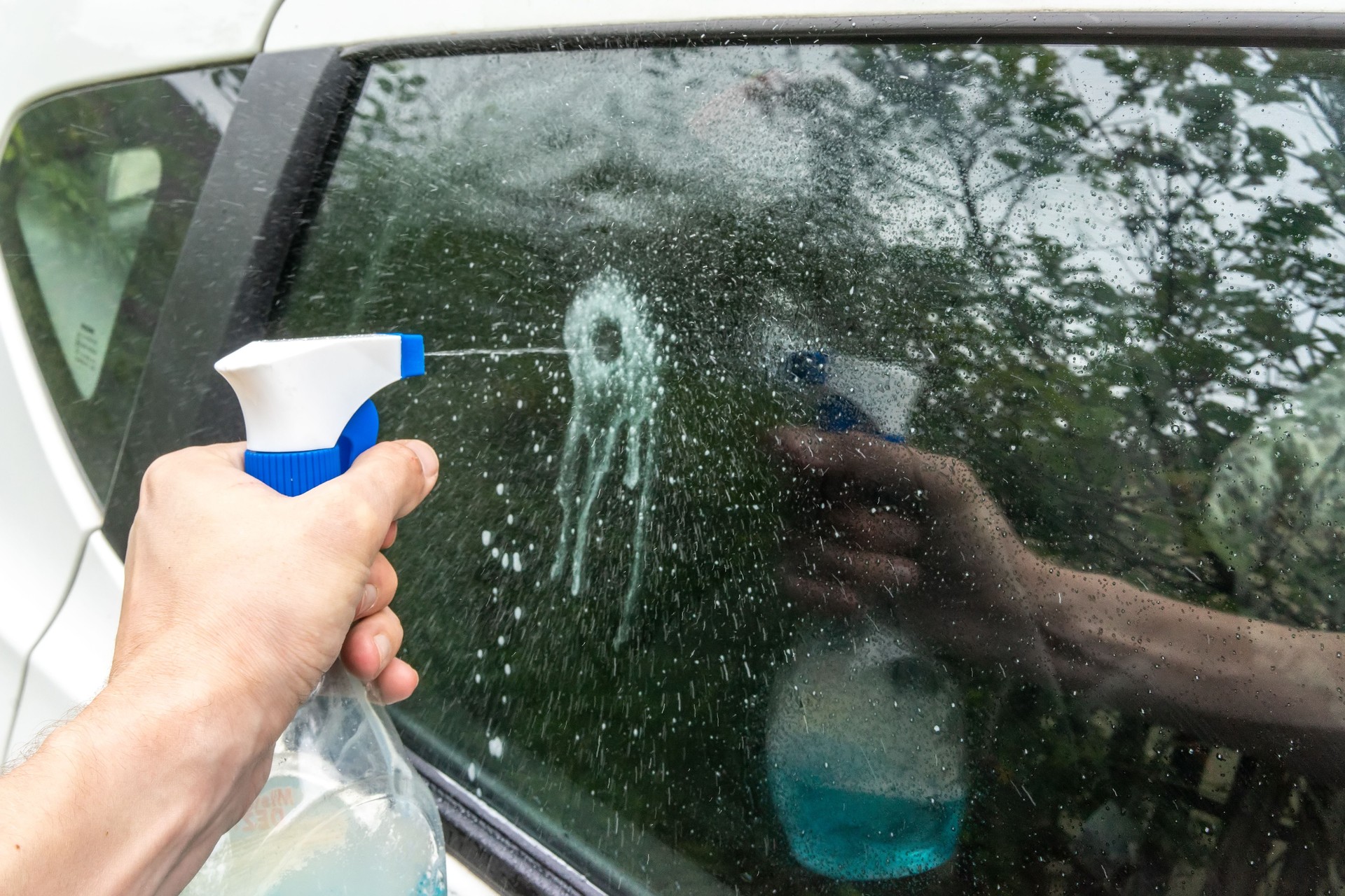 A hand holding a plastic spray bottle splashes onto a car's glass.